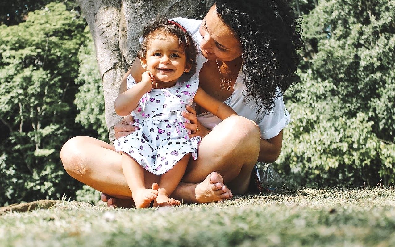 A woman with curly dark hair sits cross-legged on the grass under a tree, holding a smiling toddler in a heart-print dress
